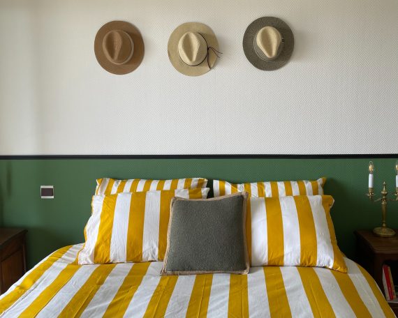 Lit avec des draps rayés jaunes et blancs, trois chapeaux accrochés au mur.