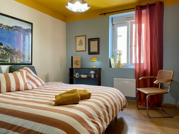 Chambre avec lit rayé, fauteuil en bois et décoration murale artistique.